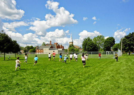 Group of children running across a field with trees and a church steeple in the background