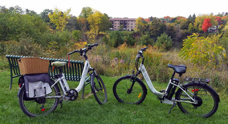 Two electric bicycles in a park, on grass, by a bench, overlooking the river. Photo credit: Green Street